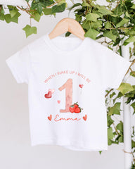 A white t-shirt with a personalized design hanging on a wooden hanger, featuring a large number one, hearts, and the text "When I wake up I will be 1 Emma".