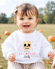 A smiling baby girl wearing a white sweater with a skull design and text, sitting in a field with pumpkins in the background.