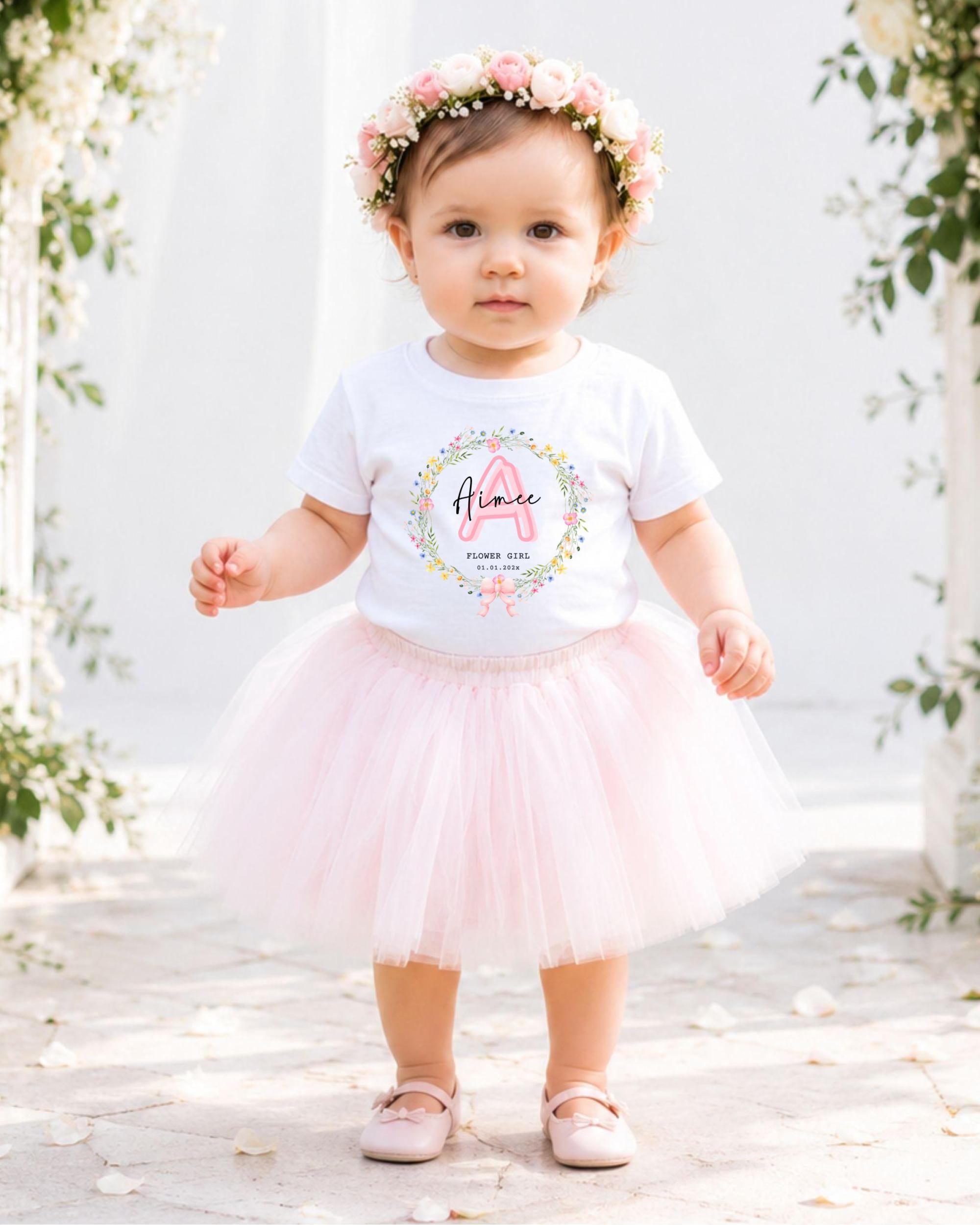 A young girl wearing a white t-shirt with a floral design and a pink tulle skirt, standing in front of a white arch with flowers.