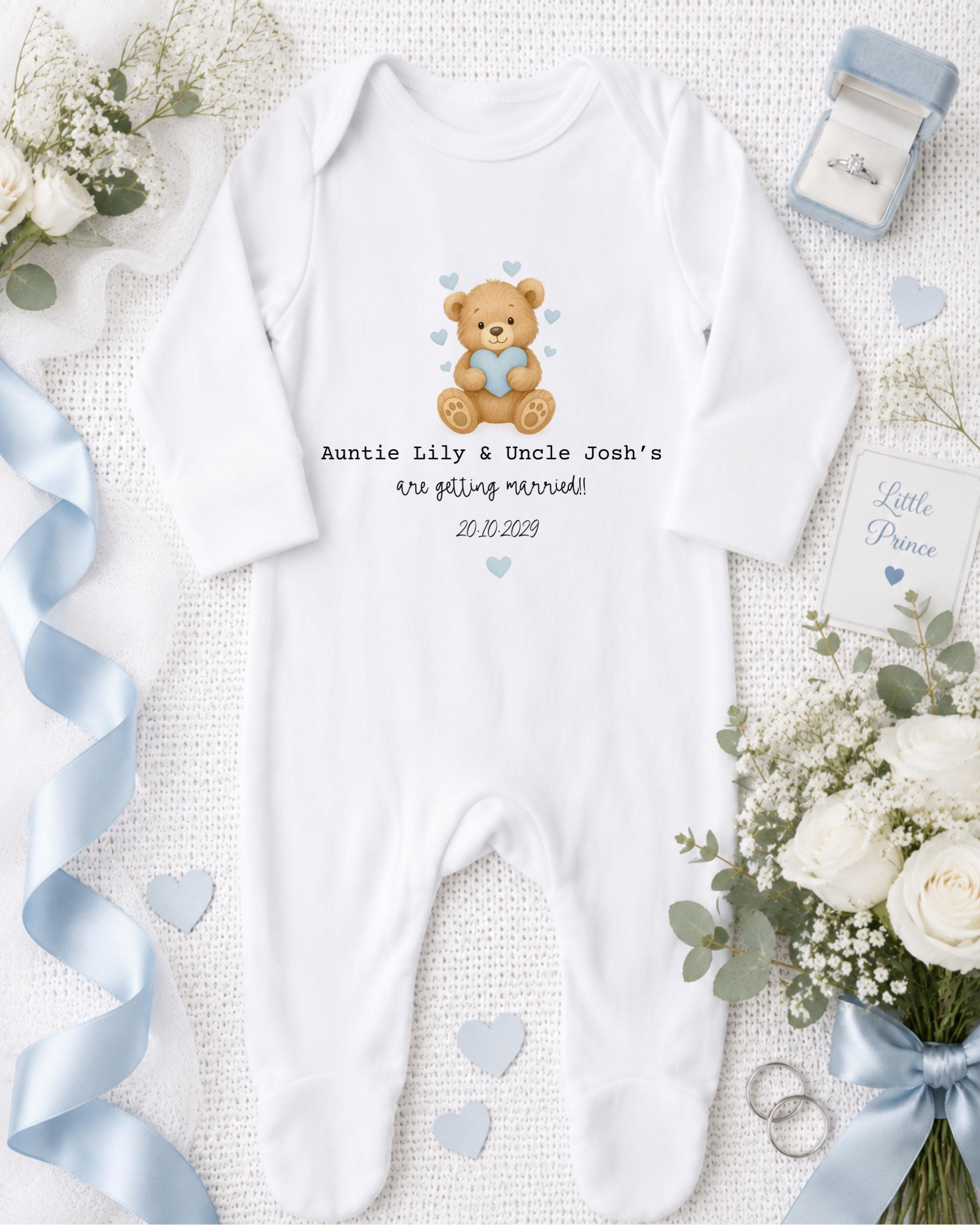 A white onesie with a teddy bear graphic and text, along with a blue ribbon, white flowers, and a ring, all arranged on a white background.