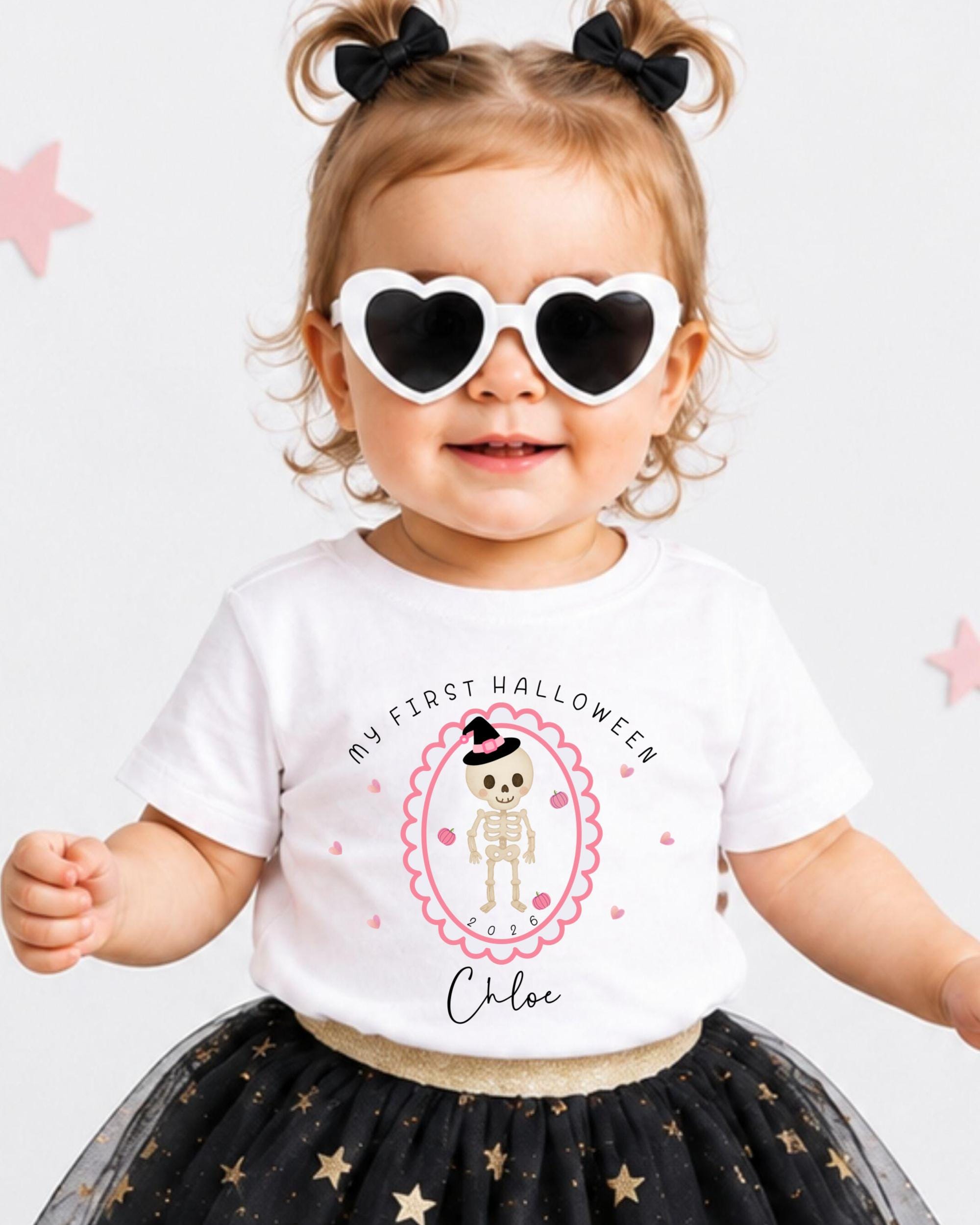 A young girl wearing a white t-shirt with a skeleton design and heart-shaped sunglasses, standing in front of pink stars.