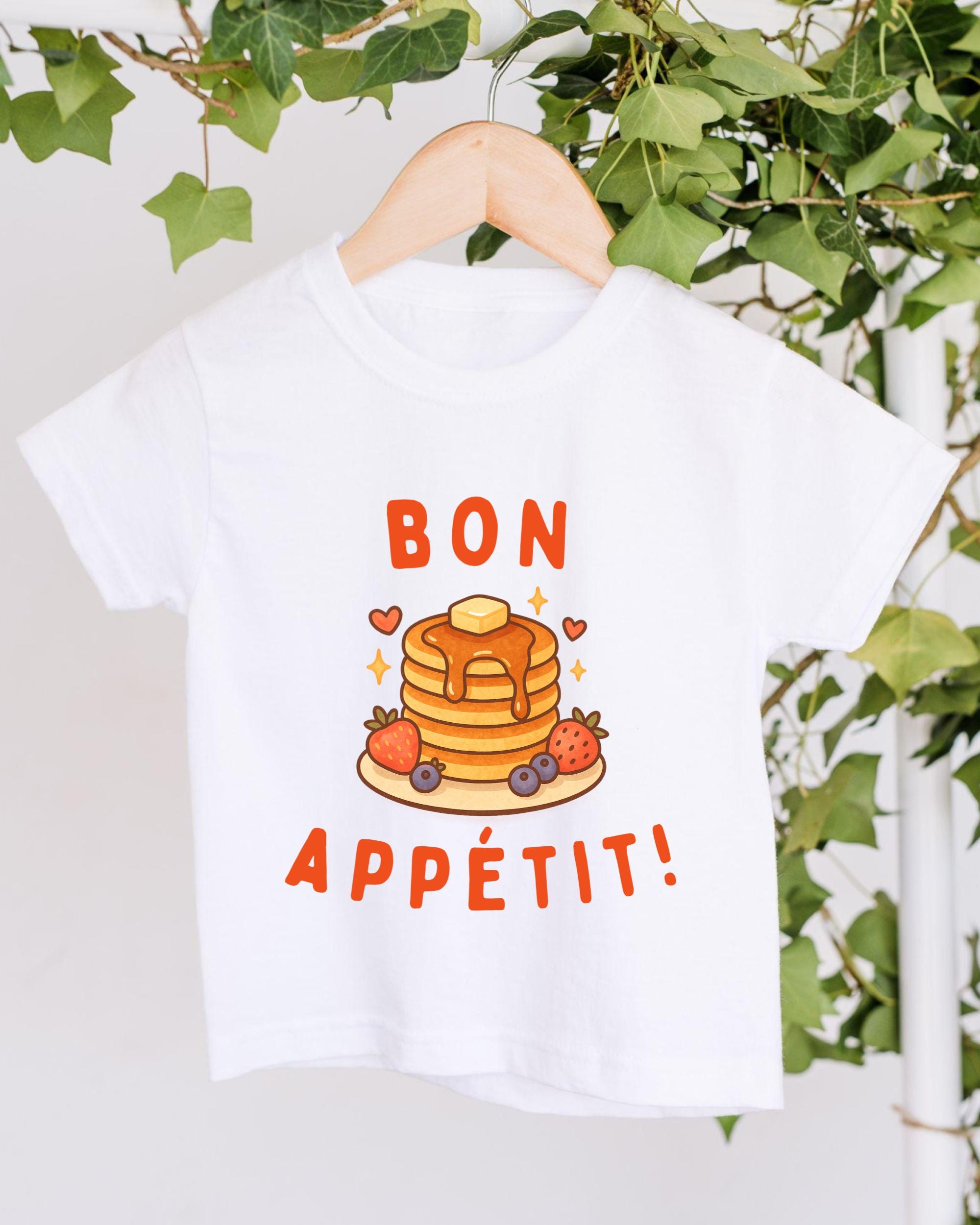 A white t-shirt with a graphic design featuring pancakes, strawberries, and the text "BON APETIT!"