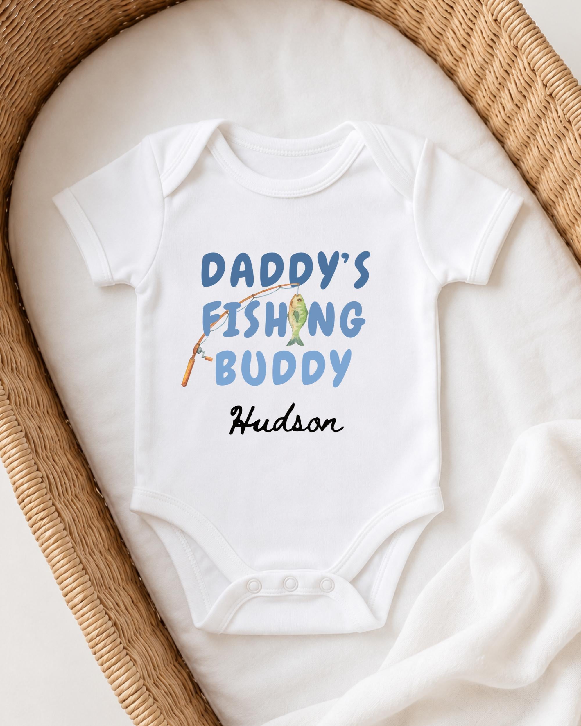 A white baby onesie with the text "DADDY'S FISHING BUDDY" printed on it, along with an illustration of a fishing rod and fish.