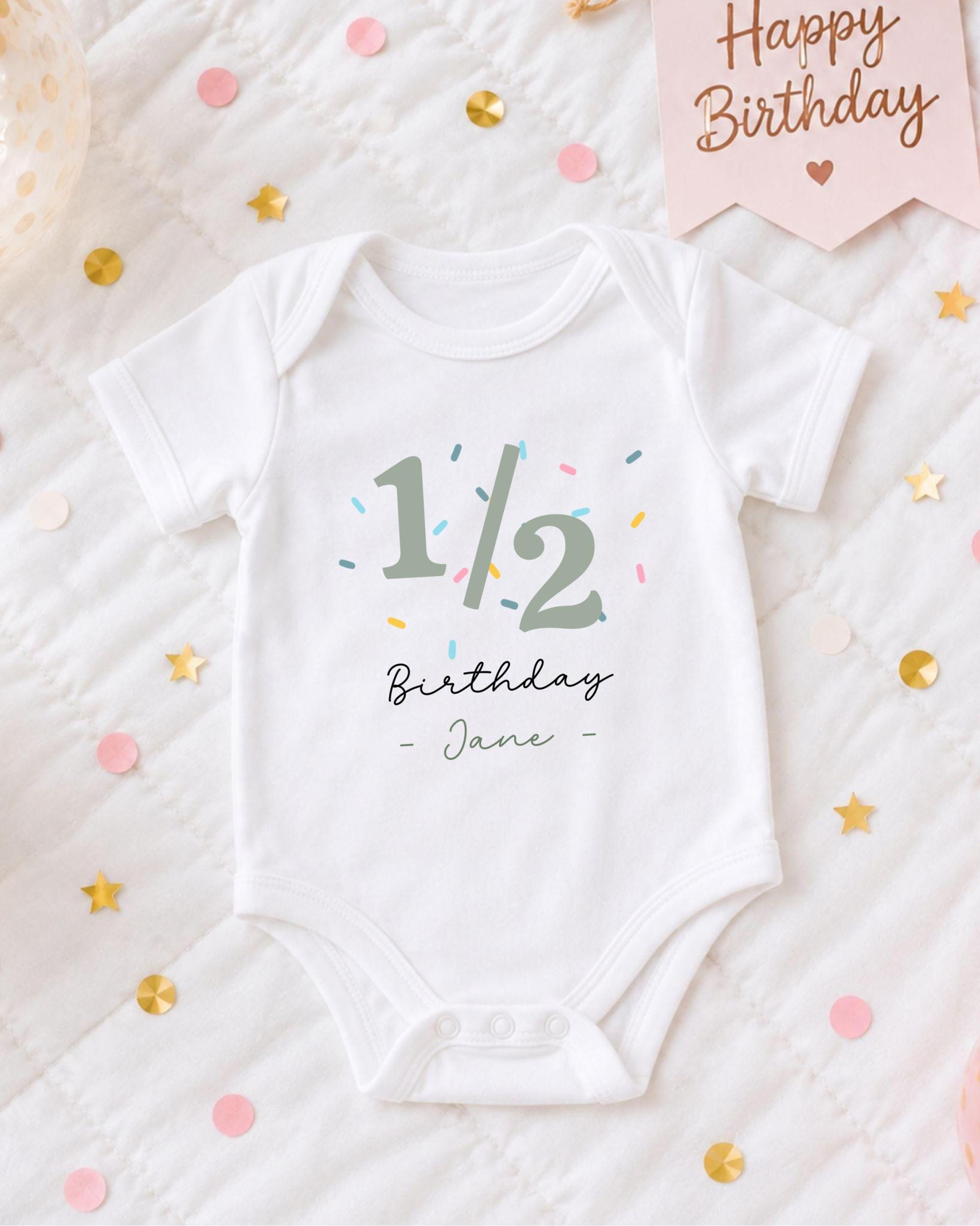 A white onesie with the text "1/2 Birthday - Jane" printed on it, along with a pink banner that says "Happy Birthday" and confetti scattered around.