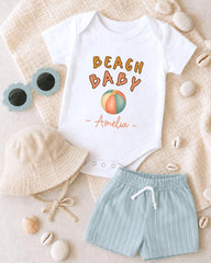A white onesie with the text "BEACH BABY" printed on it, along with a bucket hat, sunglasses, and a pair of shorts, all laid out on a beige surface.