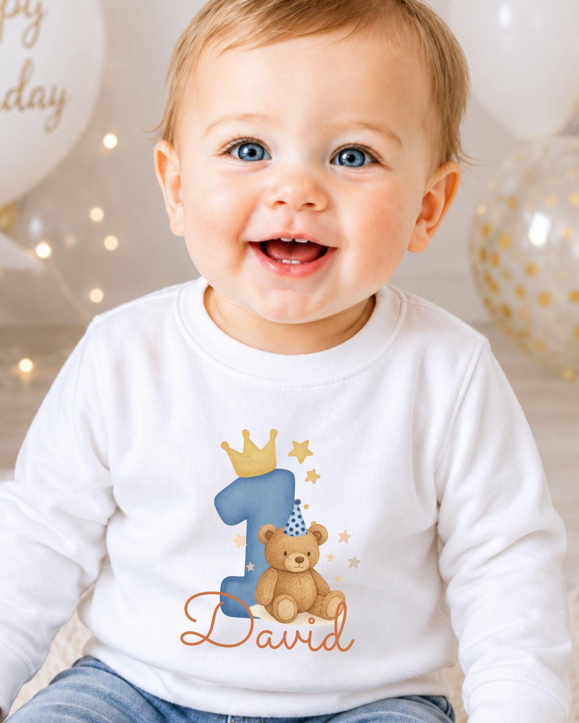 A smiling baby boy wearing a white shirt with a teddy bear and a number one, celebrating his first birthday.