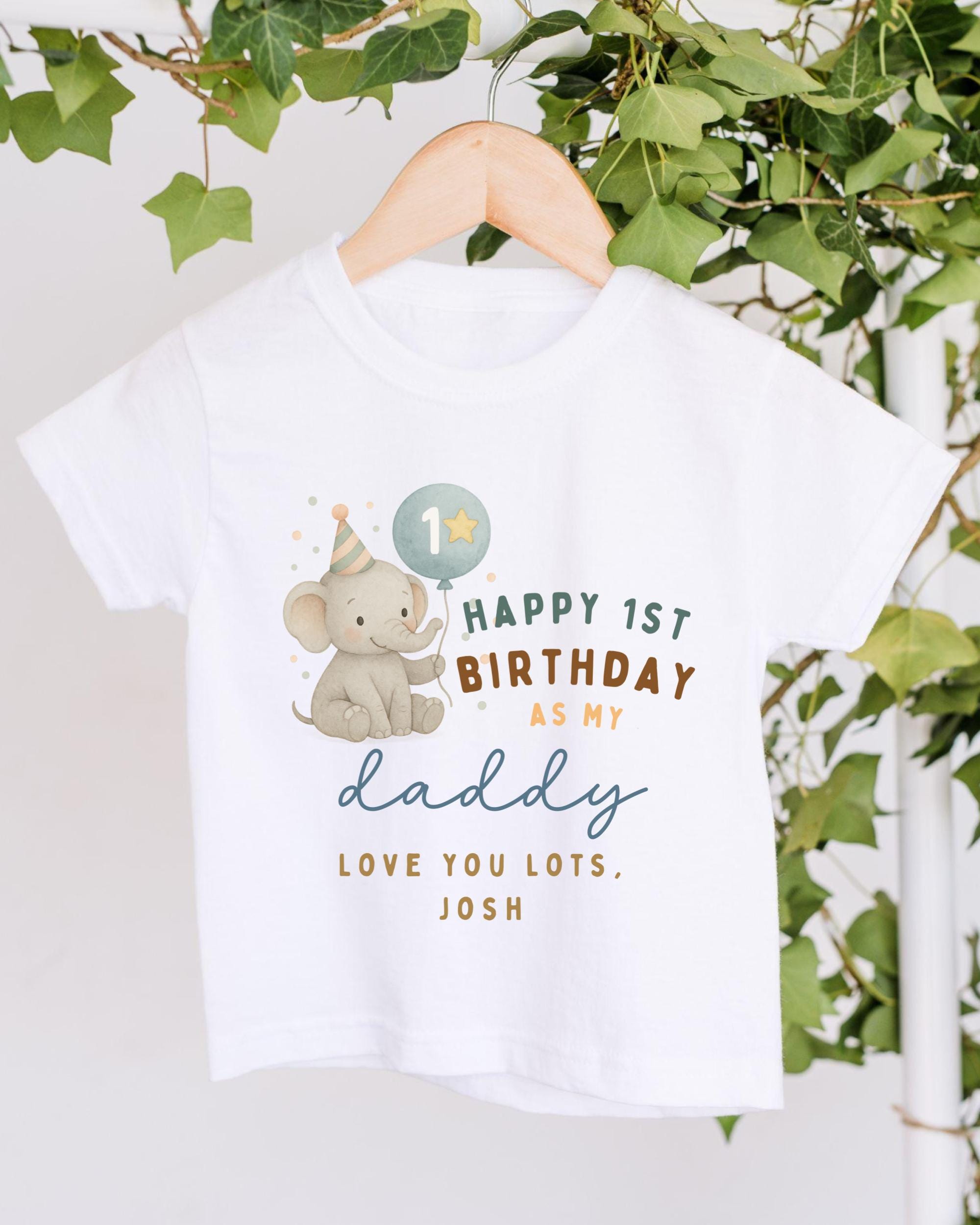 A white t-shirt with a graphic design featuring a baby elephant wearing a party hat and the text "Happy 1st Birthday as my daddy, love you lots, Josh" printed on it.