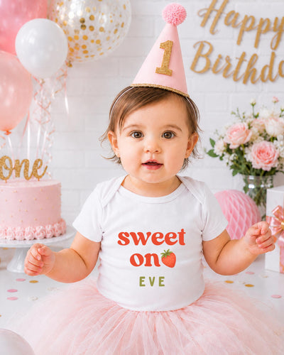 A young girl wearing a birthday hat and a white t-shirt with the text "Sweet on Eve" is posing in front of a birthday cake and balloons.