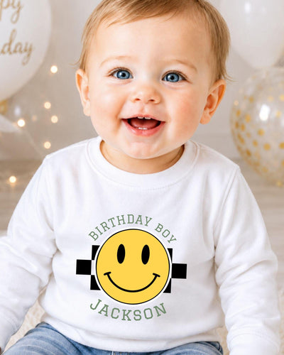 A smiling baby boy wearing a white shirt with a yellow smiley face and text that says "Birthday Boy Jackson".
