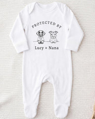 A white onesie with a graphic design featuring two cartoon dogs and the text "PROTECTED BY Lucy + Nana" printed on it.