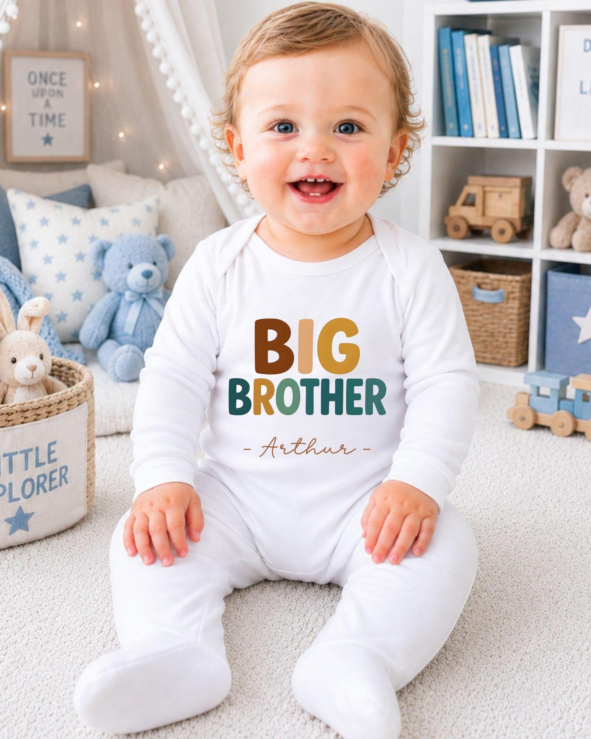 A smiling baby boy wearing a white onesie with the text "BIG BROTHER Arthur" sits on the floor in a nursery setting.
