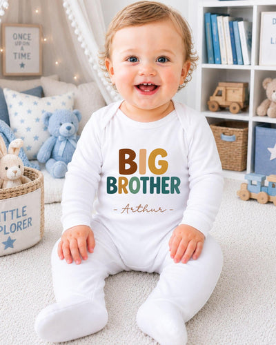 A smiling baby boy wearing a white bodysuit with the text "BIG BROTHER Arthur" sits on the floor in a nursery setting.