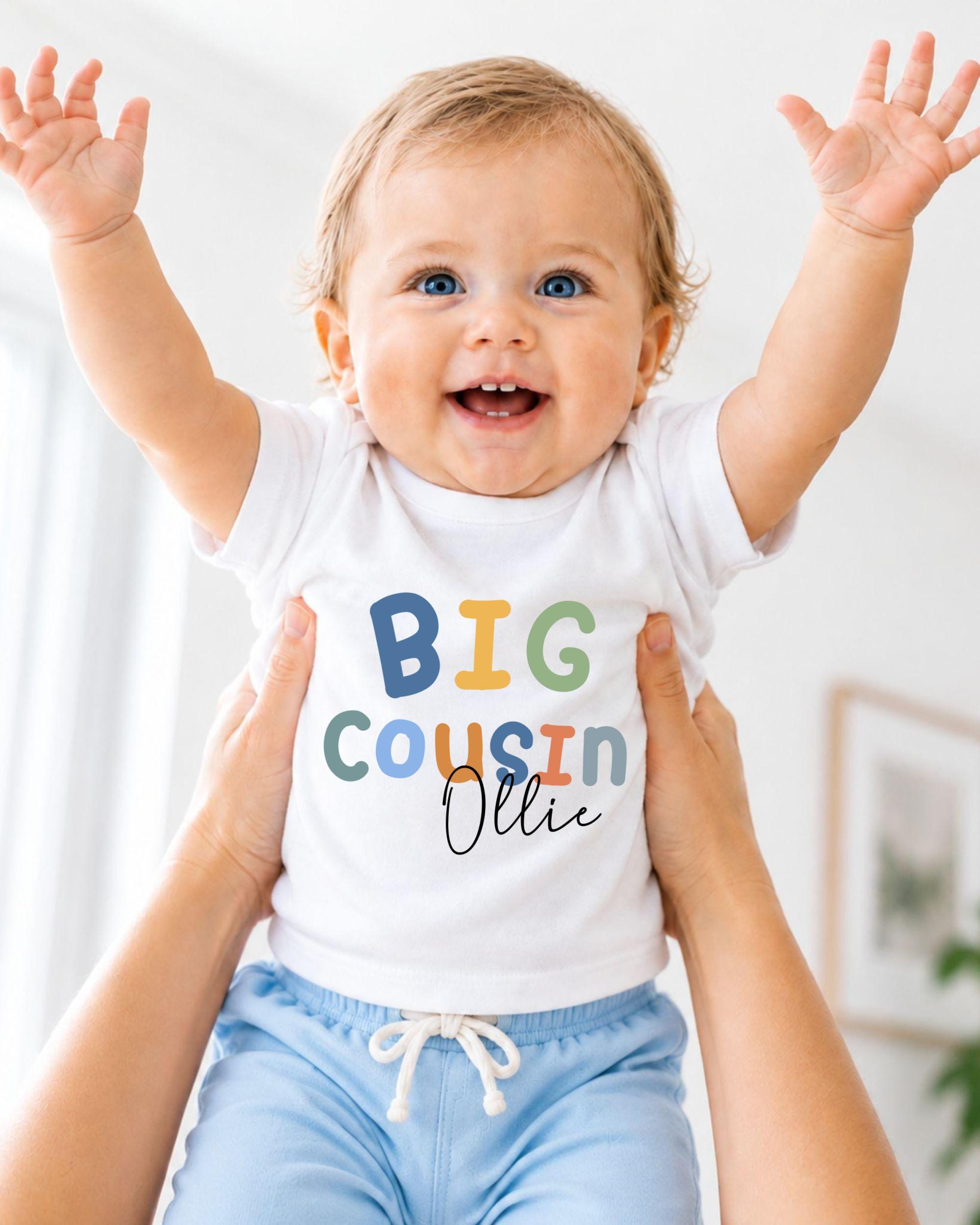 A smiling baby wearing a white t-shirt with the text "BIG COUSIN OLLIE" is being held up by someone's arms.