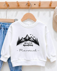 A white sweatshirt with a mountain landscape design and the text "Take me to the mountains" printed on it, hanging on a wooden hanger.