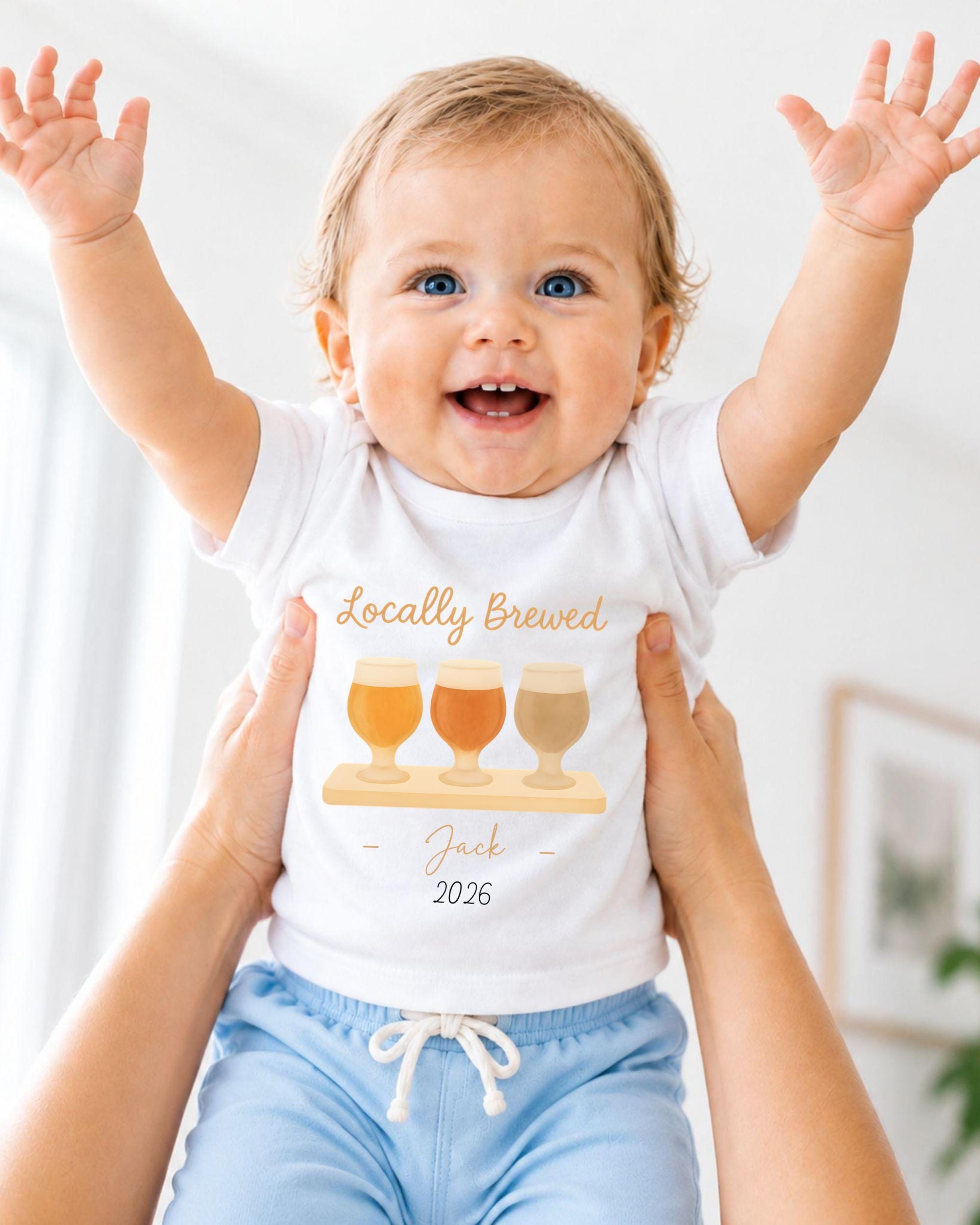 A smiling baby wearing a white shirt with a graphic of three beer glasses and the text "Locally Brewed Jack 2026" is being held up by someone's arms.