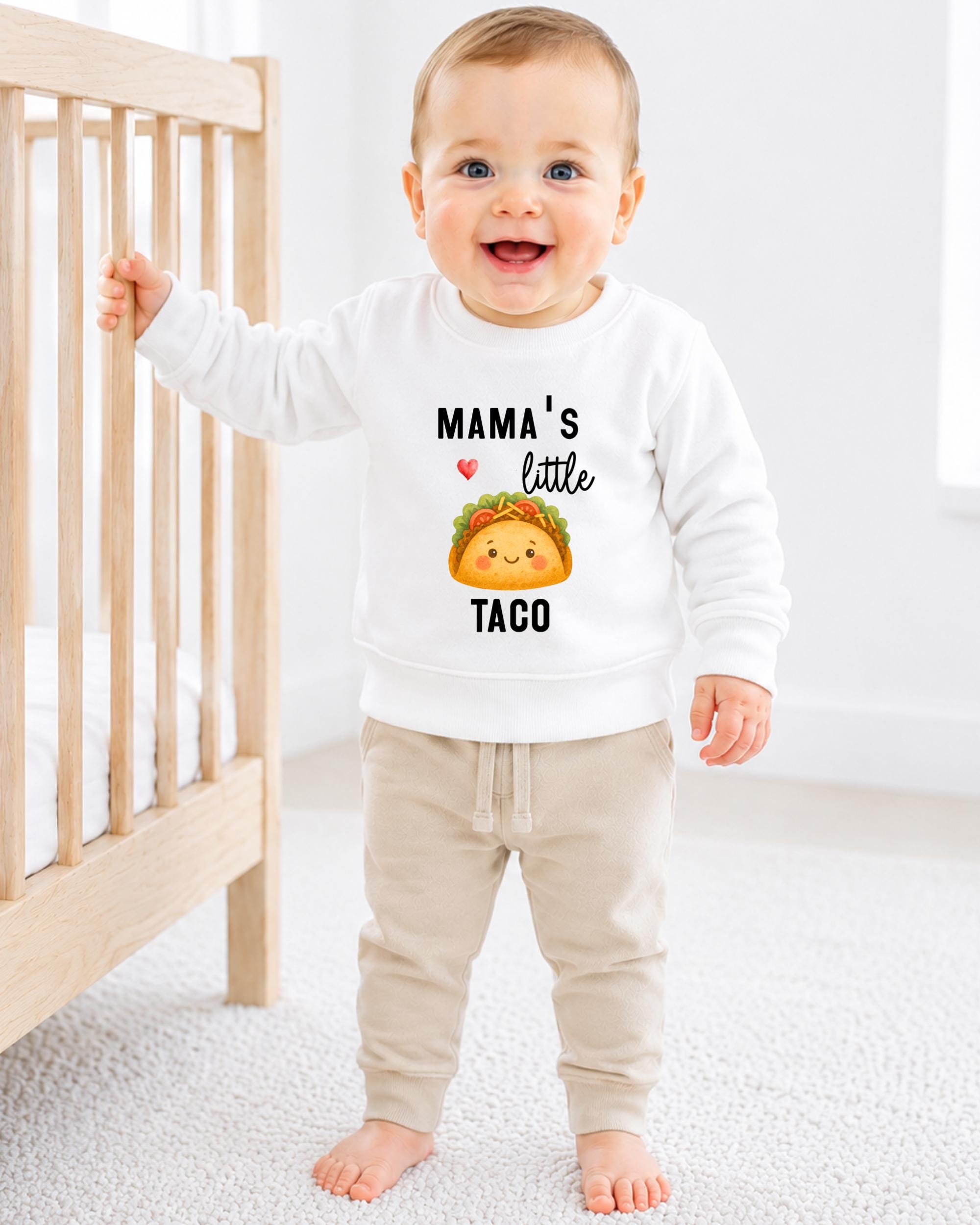 A smiling baby boy wearing a white sweatshirt with a cartoon taco graphic and the text "Mama's little taco" printed on it.