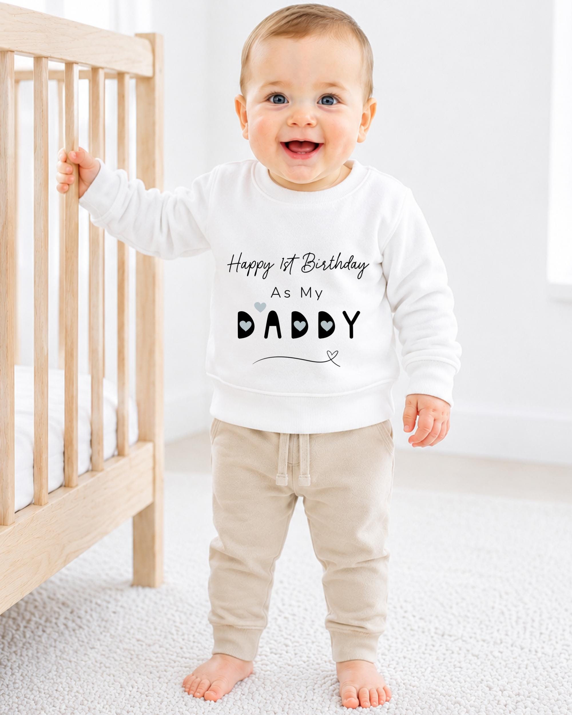 A smiling baby boy wearing a white sweatshirt with the text "Happy 1st Birthday as my Daddy" printed on it.