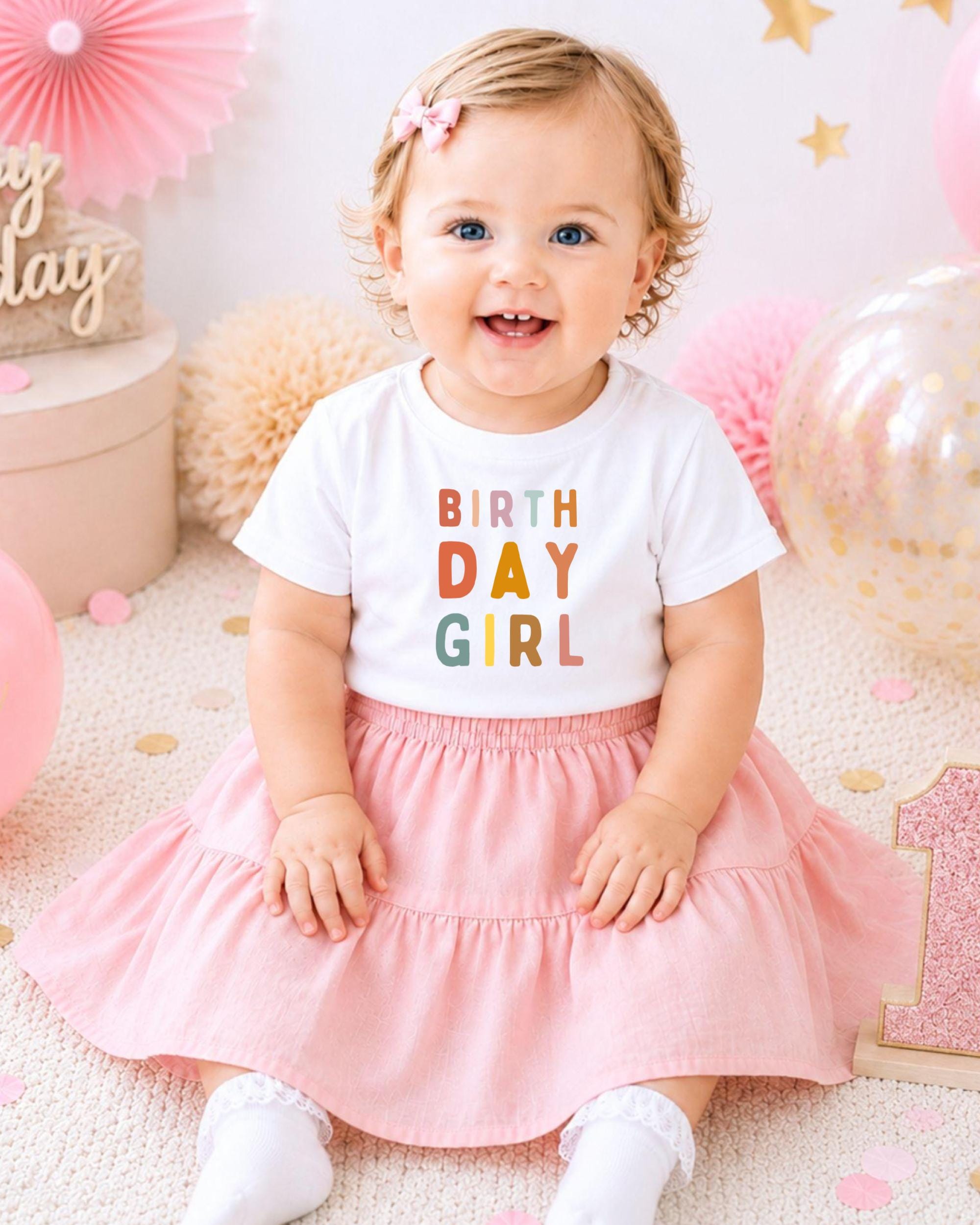 A smiling baby girl wearing a white t-shirt with the text "BIRTH DAY GIRL" and a pink skirt, sitting on a pink and white background with balloons and confetti.