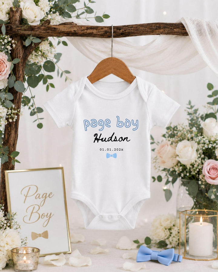 A baby boy's white bodysuit with the text "Page Boy Hudson" printed on it, hanging on a wooden hanger. The bodysuit is displayed in a decorative setting with floral arrangements, candles, and a framed sign.