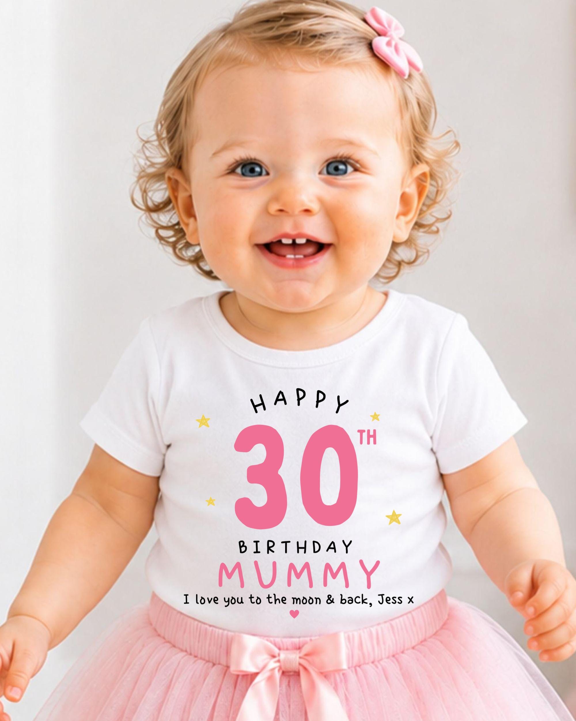 A smiling baby girl wearing a white t-shirt with a birthday message and a pink bow in her hair.