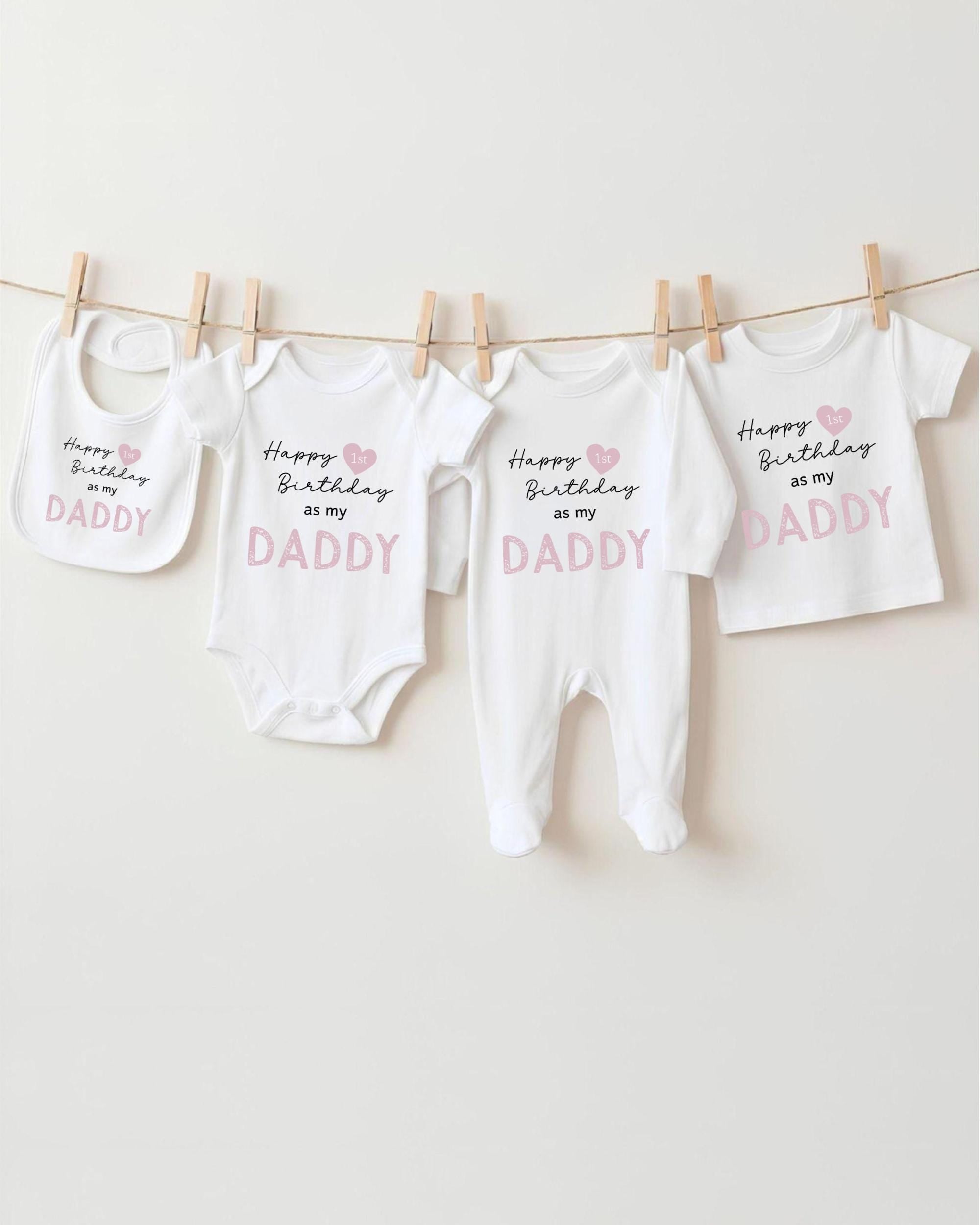 A collection of four white baby clothes, including onesies and bibs, hanging on a clothesline. Each piece of clothing has a message printed on it, likely related to a birthday celebration.