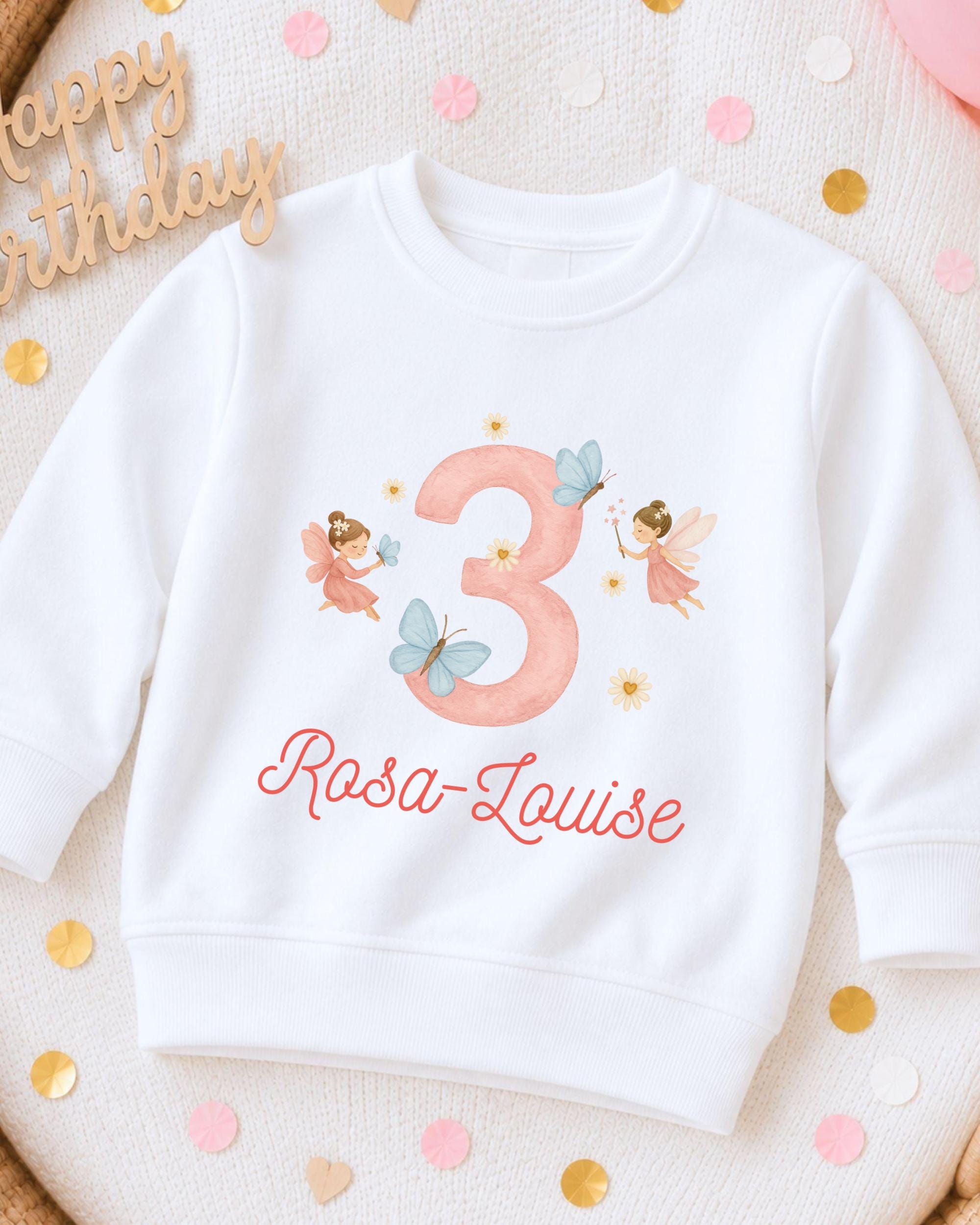 A white sweatshirt with a pink number 3 and a fairy design, along with the text "Rosa-Louise" printed on it.