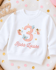 A white sweatshirt with a pink number 3 and a fairy design, along with the text "Rosa-Louise" printed on it.