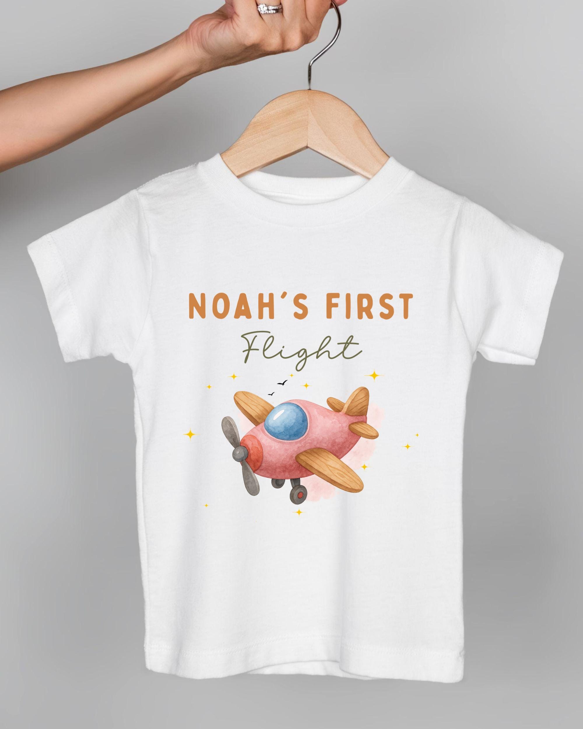 A white t-shirt with a pink airplane graphic and the text "Noah's first flight" printed on it.