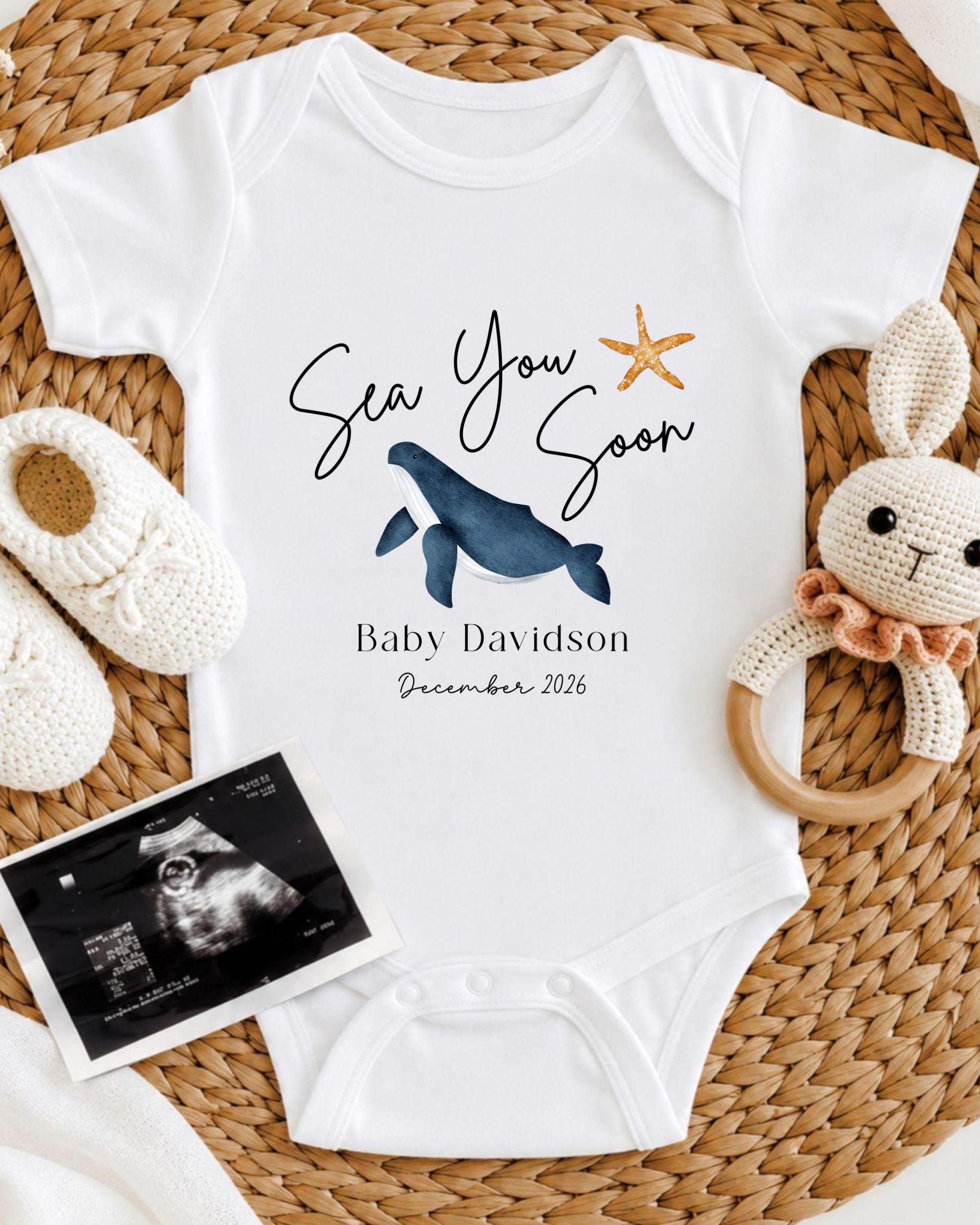 a baby announcement, including a white onesie with a blue whale and starfish design, a crocheted bunny toy, and a baby's firstborn announcement.
