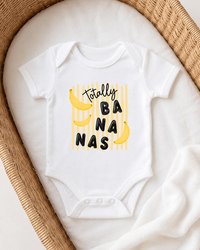 A white onesie with a playful banana graphic and the text "Totally BANANA" printed on it, laid out on a white blanket.
