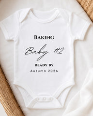 A white baby onesie with the text "BAKING Baby #2 READY BY Autumn 2026" printed on it.