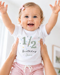 A smiling baby girl wearing a white t-shirt with the number 1/2 and the text "Birthday Jane" printed on it, being held up by someone's arms.