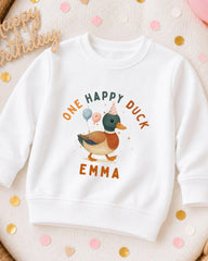 A white sweatshirt with a cartoon duck and the text "One Happy Duck Emma" printed on it, surrounded by confetti.