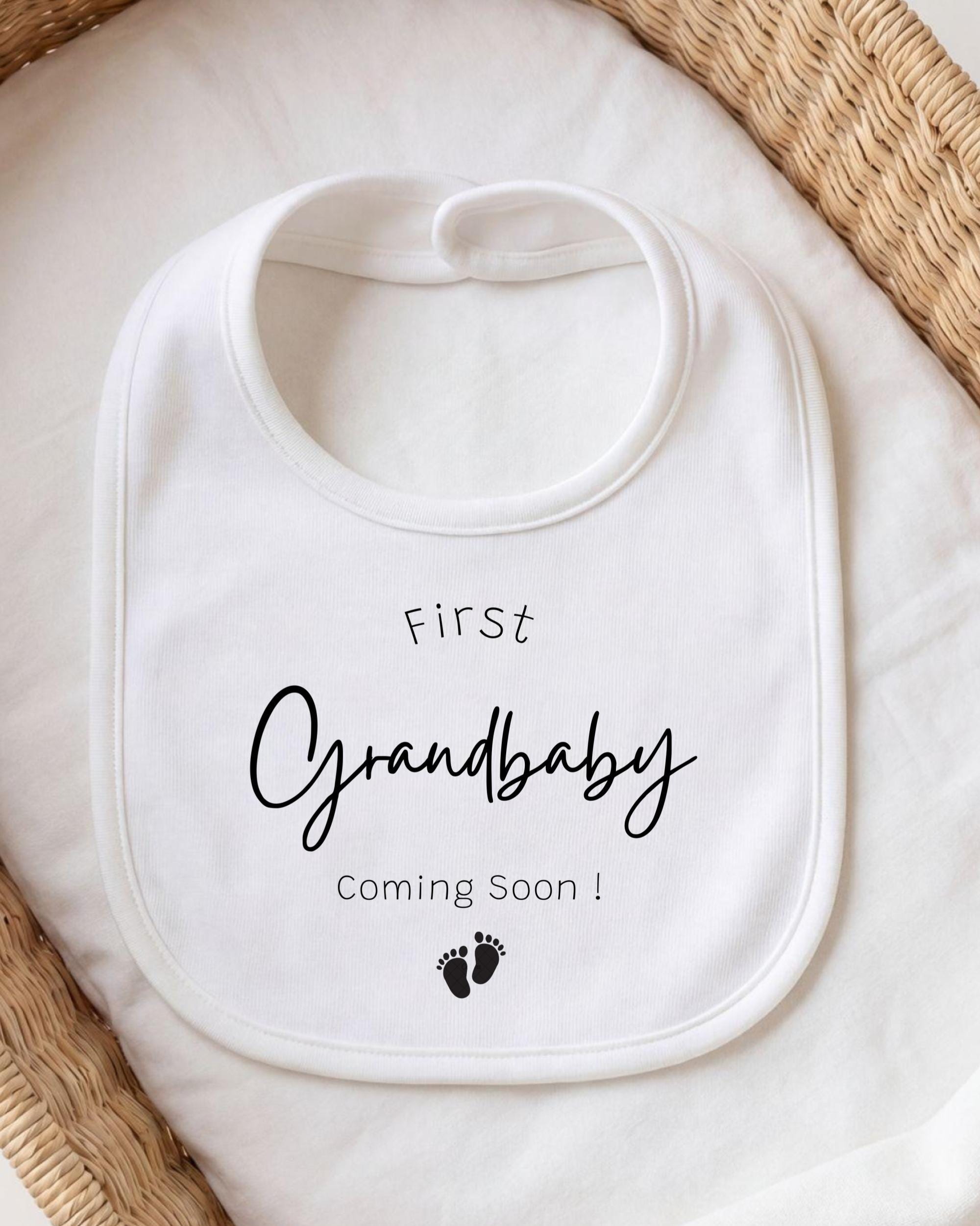 A white bib with the text "First Grandbaby Coming Soon!" printed on it, placed on a white cloth.