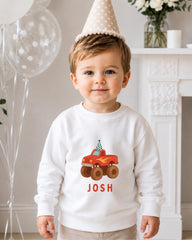 A young boy wearing a white sweater with a red truck and the name " Josh" printed on it, standing in front of a fireplace with balloons in the background.