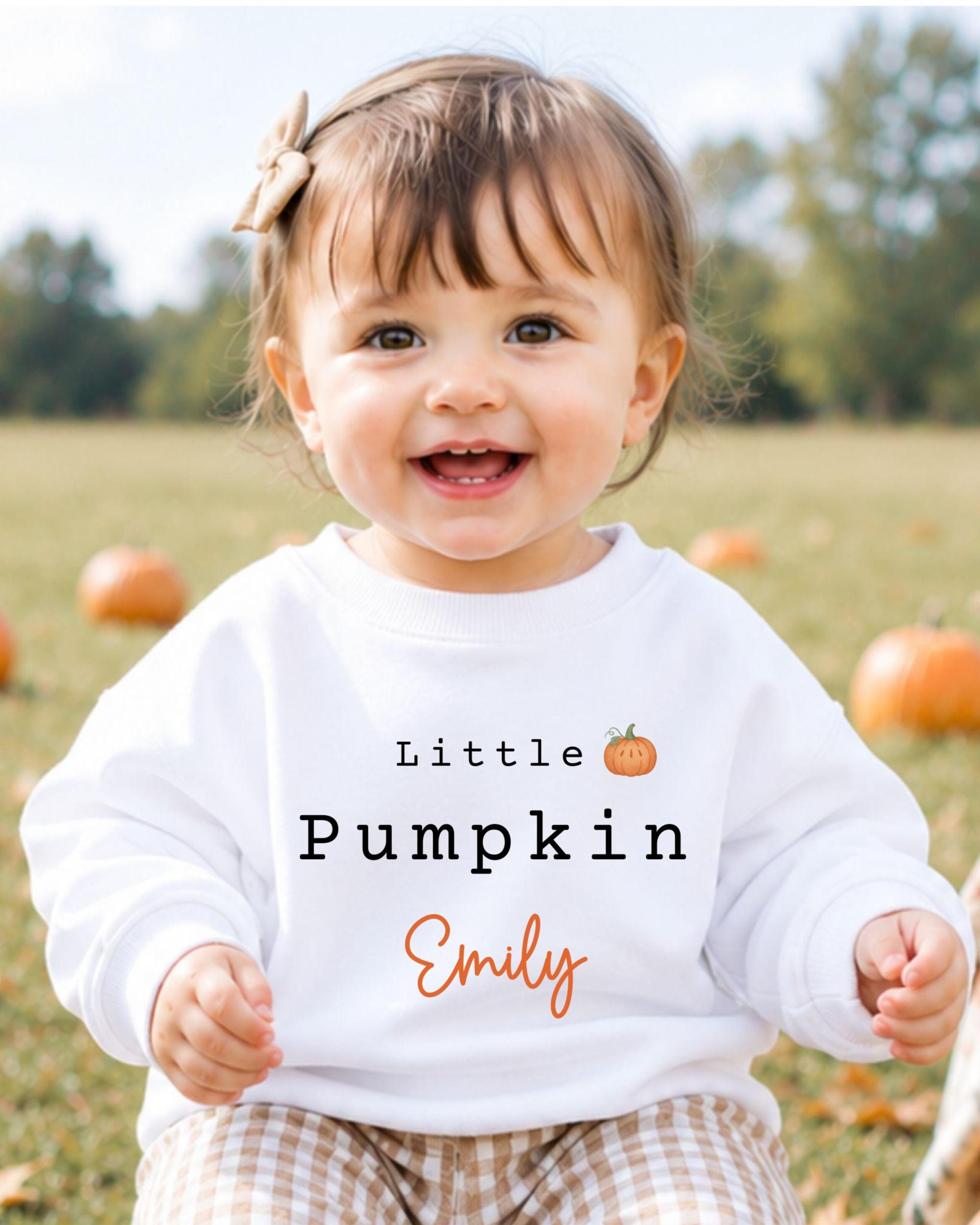 A smiling baby girl wearing a white sweatshirt with the text "Little Pumpkin Emily" printed on it, sitting in a field with pumpkins in the background.