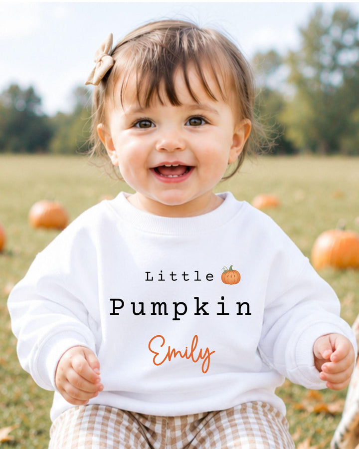 A smiling baby girl wearing a white sweatshirt with the text "Little Pumpkin Emily" printed on it, sitting in a field with pumpkins in the background.