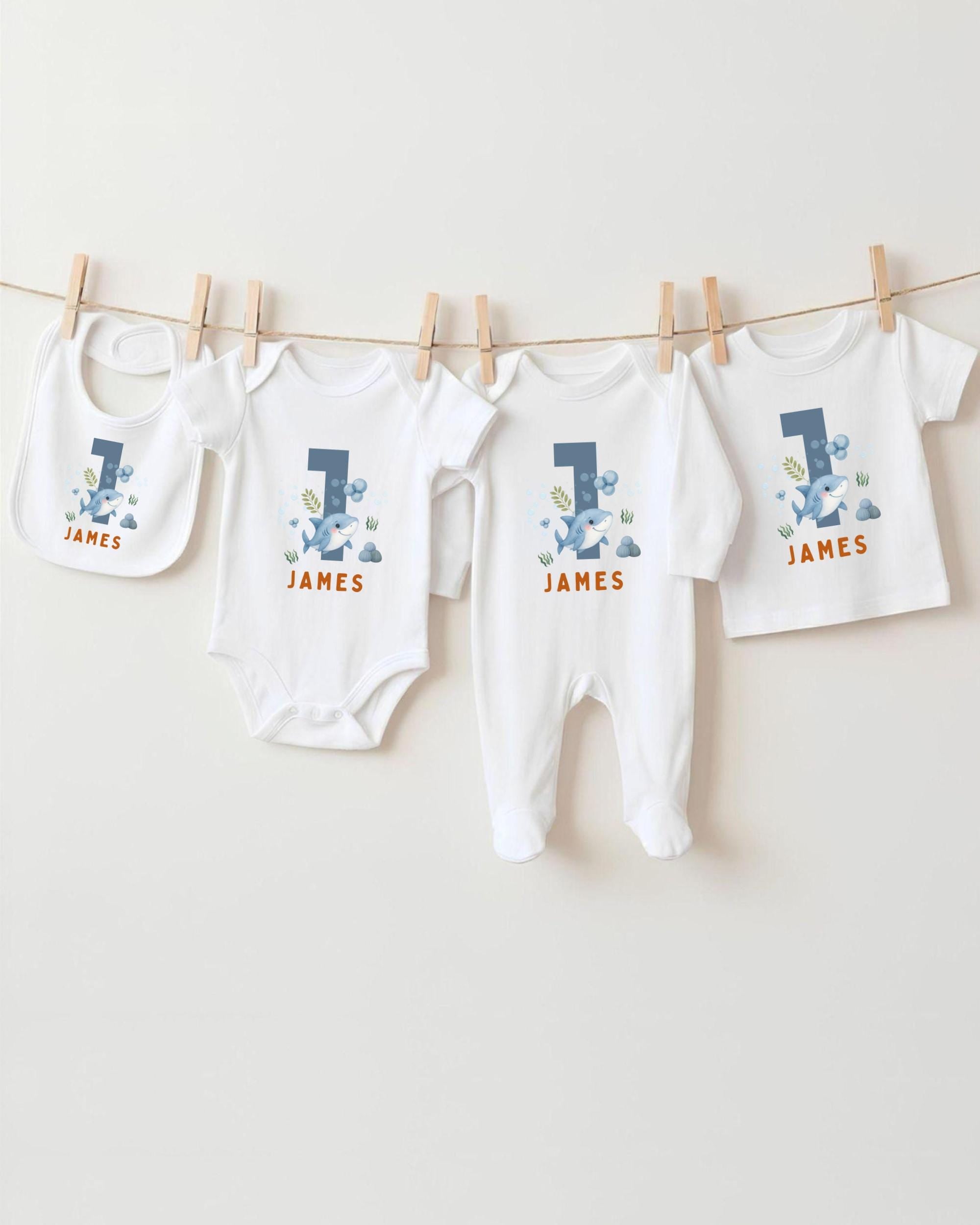 A collection of white baby clothes, including onesies and pants, with the name "James" printed on them. The clothes are hanging on a clothesline, with the first two items being bibs and the third one being a regular-sized shirt.