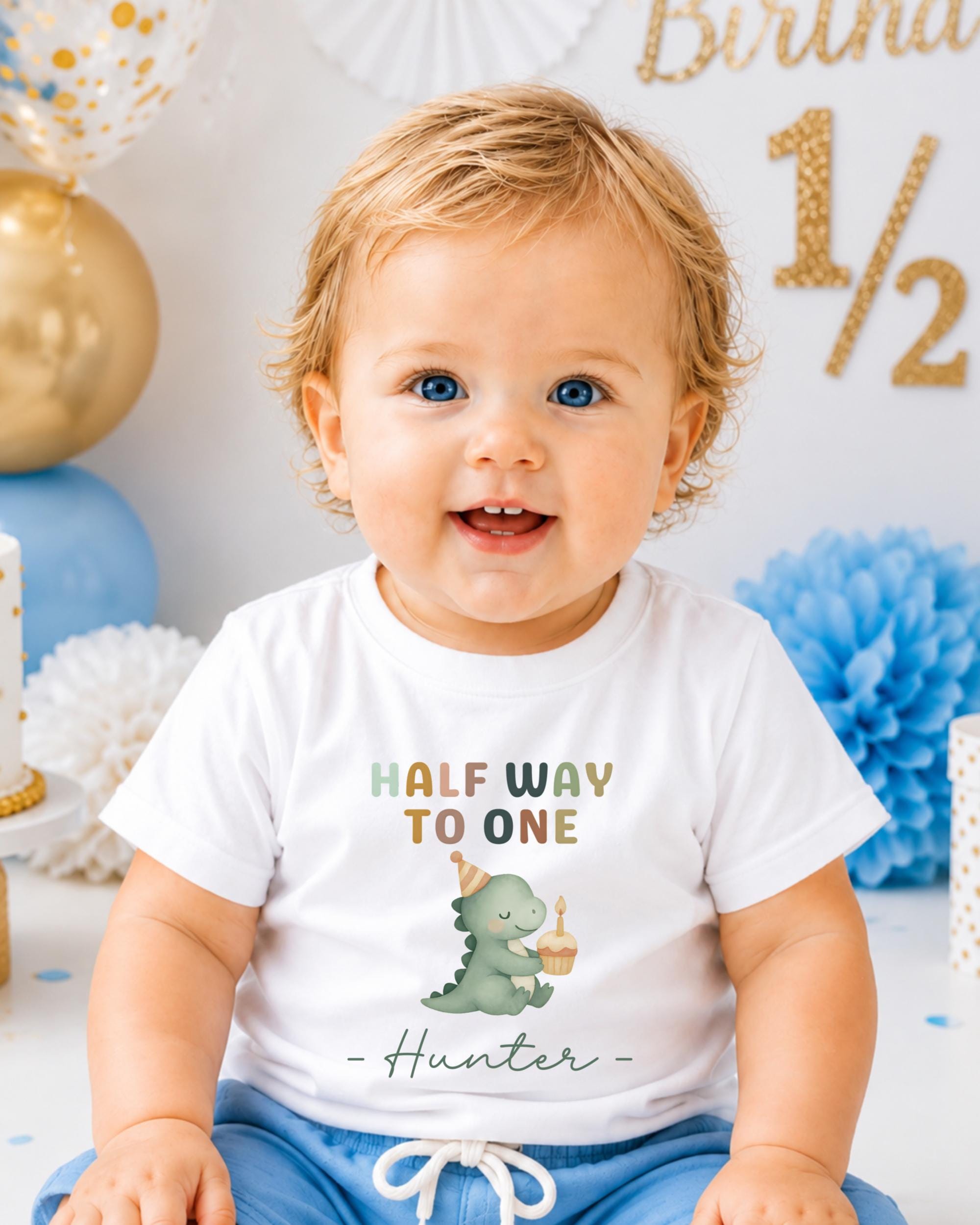 A smiling baby boy wearing a white t-shirt with a dinosaur graphic and text that says "Half way to one Hunter".