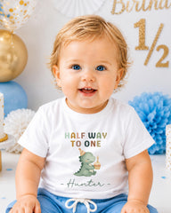 A smiling baby boy wearing a white t-shirt with a dinosaur graphic and text that says "Half way to one Hunter".