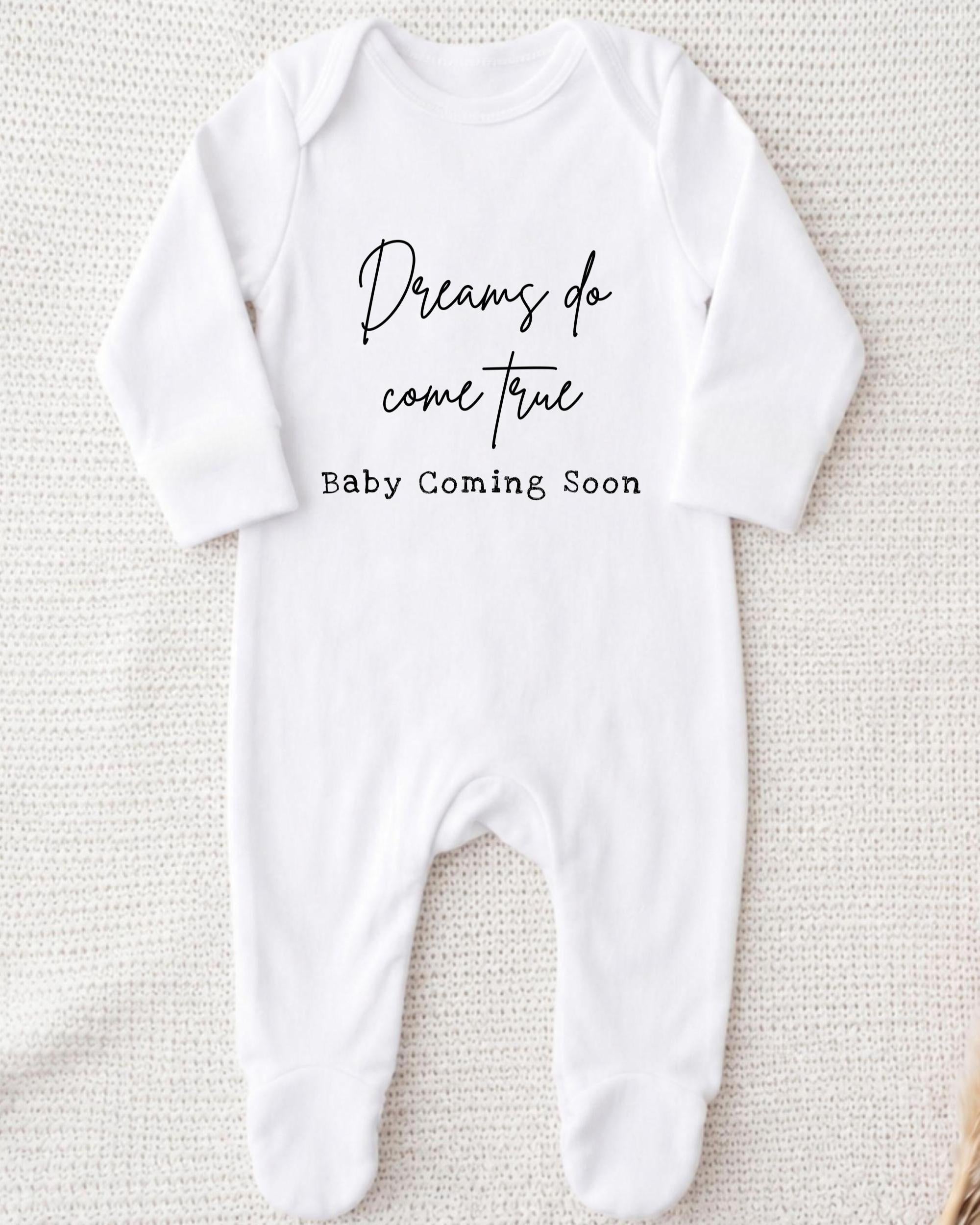 A white onesie with the text "Dreams do come true Baby Coming Soon" printed on it.
