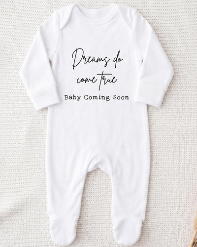 A white bodysuit with the text "Dreams do come true Baby Coming Soon" printed on it.
