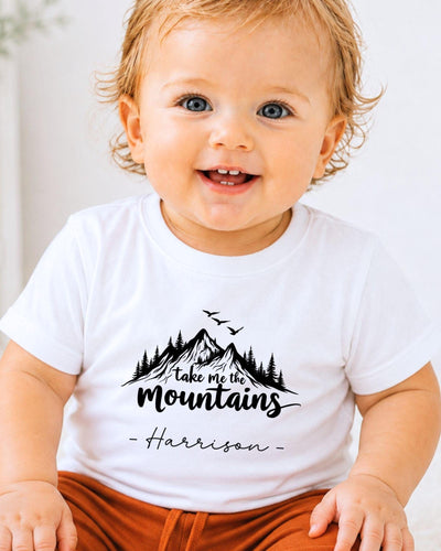 A smiling baby wearing a white t-shirt with a mountain graphic and the text "Take me the mountains" printed on it.