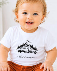 A smiling baby wearing a white t-shirt with a mountain graphic and the text "Take me the mountains" printed on it.