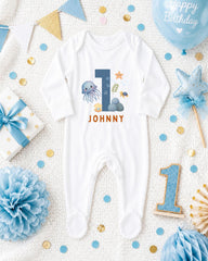 A white onesie with the number 1 and ocean-themed designs, surrounded by birthday decorations such as a blue balloon, blue and gold confetti, and a blue and white polka dot gift box.