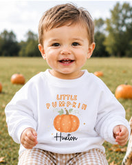 A smiling baby boy wearing a white sweatshirt with a pumpkin graphic and the text "Little Pumpkin" is sitting in a field with pumpkins in the background.
