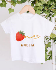 A white t-shirt with a red strawberry graphic and the name "Amelia" printed on it, hanging on a wooden hanger.