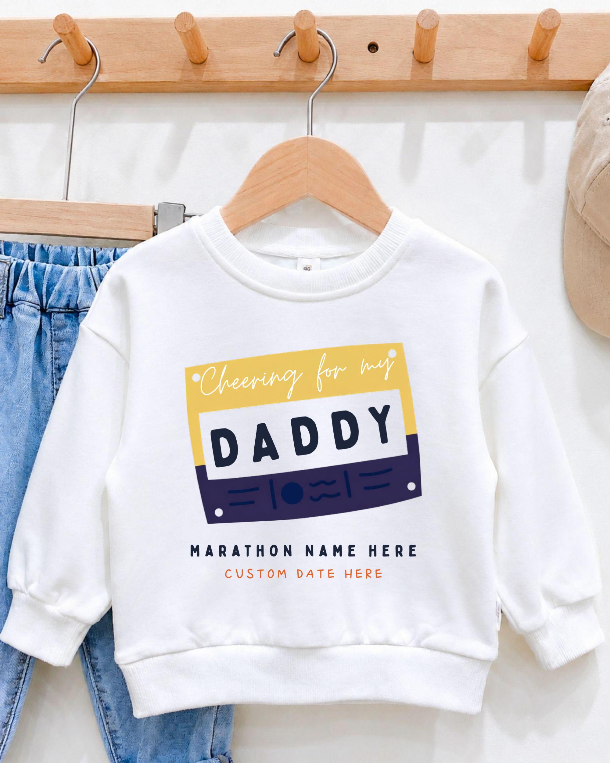 A white sweatshirt with a graphic design featuring the text "Cheering for my DADDY" and "MARCHON NAME HERE" printed on it, along with a custom date "CUSTOM DATE HERE".