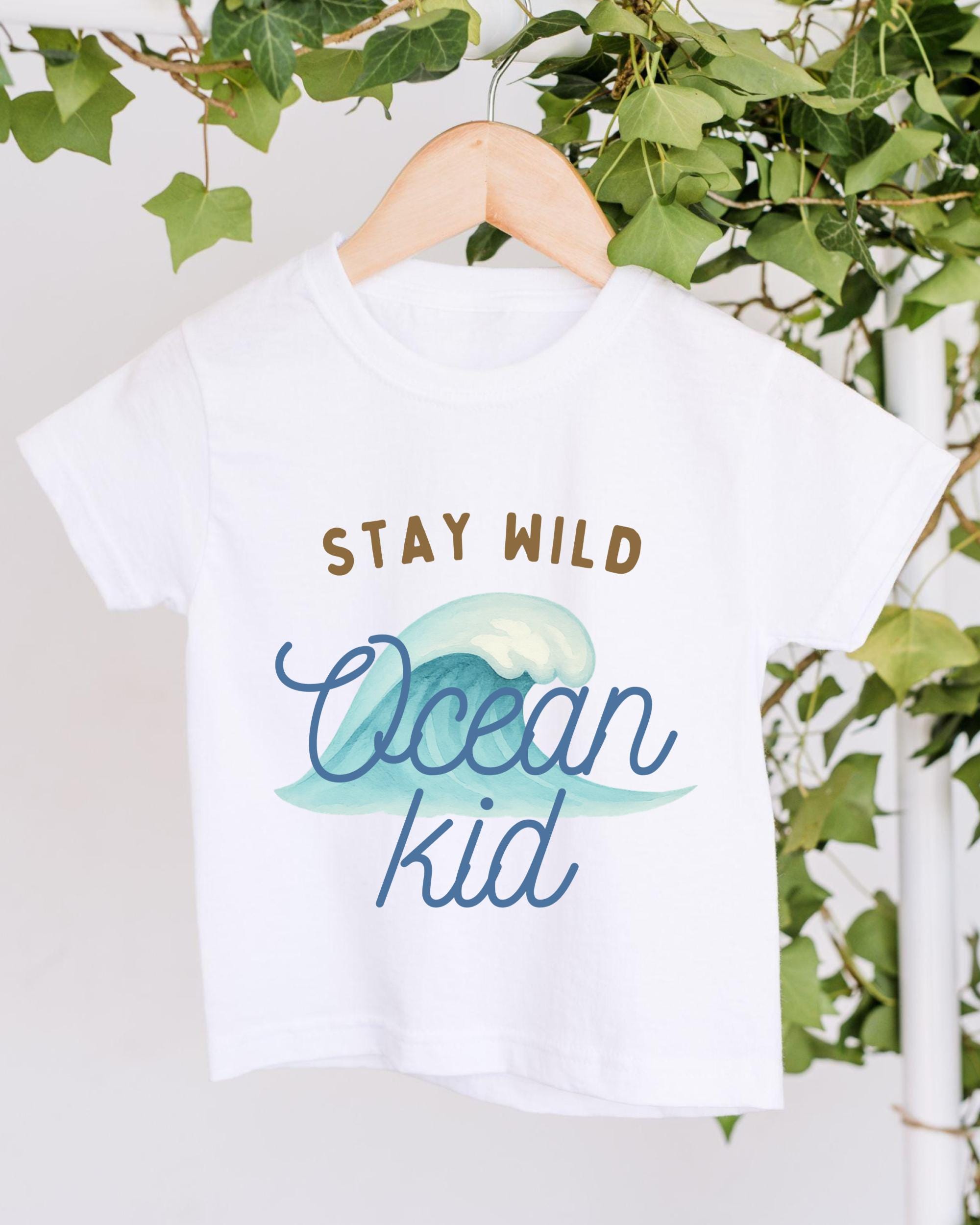 A white t-shirt with the text "STAY WILD Ocean kid" printed on it, hanging on a wooden hanger.