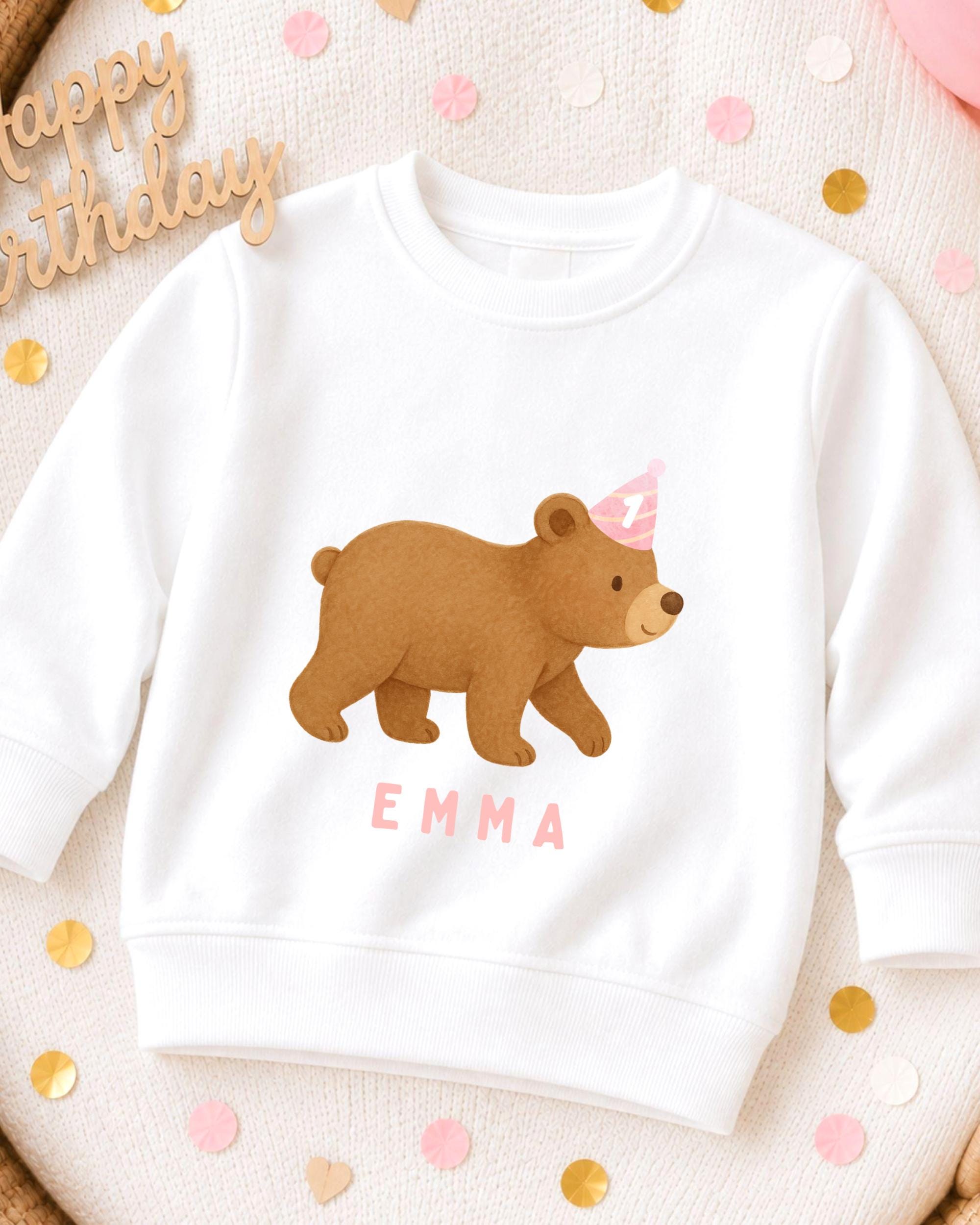 A white sweatshirt with a cartoon bear wearing a birthday hat and the name "Emma" printed on it, surrounded by confetti.